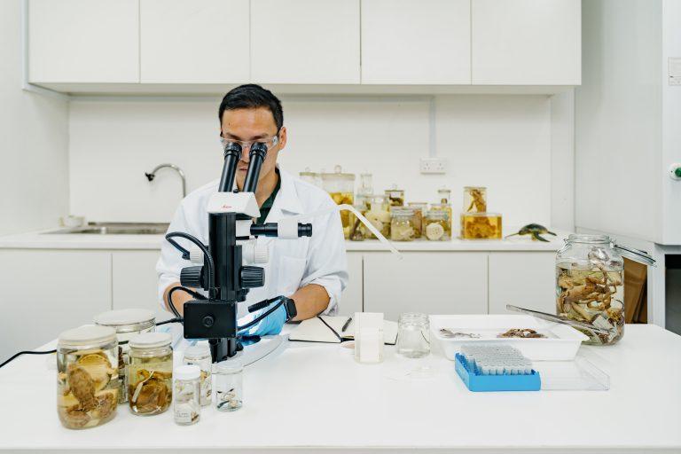 A museum staff in a white lab coat peering into a microscope with jars of specimens and lab equipment surrounding him.